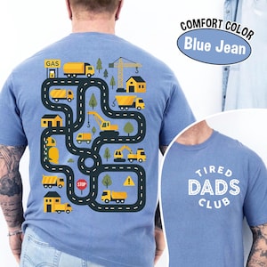 May include: Blue jean comfort color t-shirt with a playful construction site design on the back. The design features yellow trucks, a crane, houses, and trees along a winding road. The front of the shirt says "Tired Dads Club".