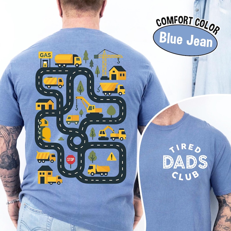 May include: Blue jean comfort color t-shirt with a playful construction site design on the back. The design features yellow trucks, a crane, houses, and trees along a winding road. The front of the shirt says "Tired Dads Club".