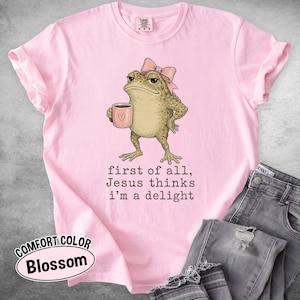 May include: A light pink t-shirt featuring a graphic of a toad wearing a pink bow and holding a pink mug with a heart. Below the toad, the text reads: first of all, Jesus thinks i'm a delight. The shirt has a crew neckline and short sleeves.