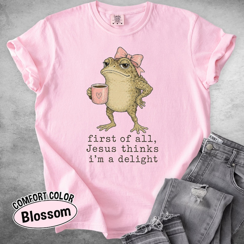 May include: A light pink t-shirt featuring a graphic of a toad wearing a pink bow and holding a pink mug with a heart. Below the toad, the text reads: first of all, Jesus thinks i'm a delight. The shirt has a crew neckline and short sleeves.