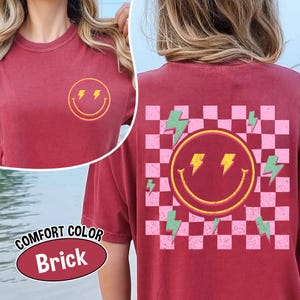 May include: Brick-colored t-shirt with a smiley face and lightning bolt design. The front features a yellow smiley face with lightning bolt eyes. The back has a larger smiley face with a checkered pink and white background and green lightning bolts.