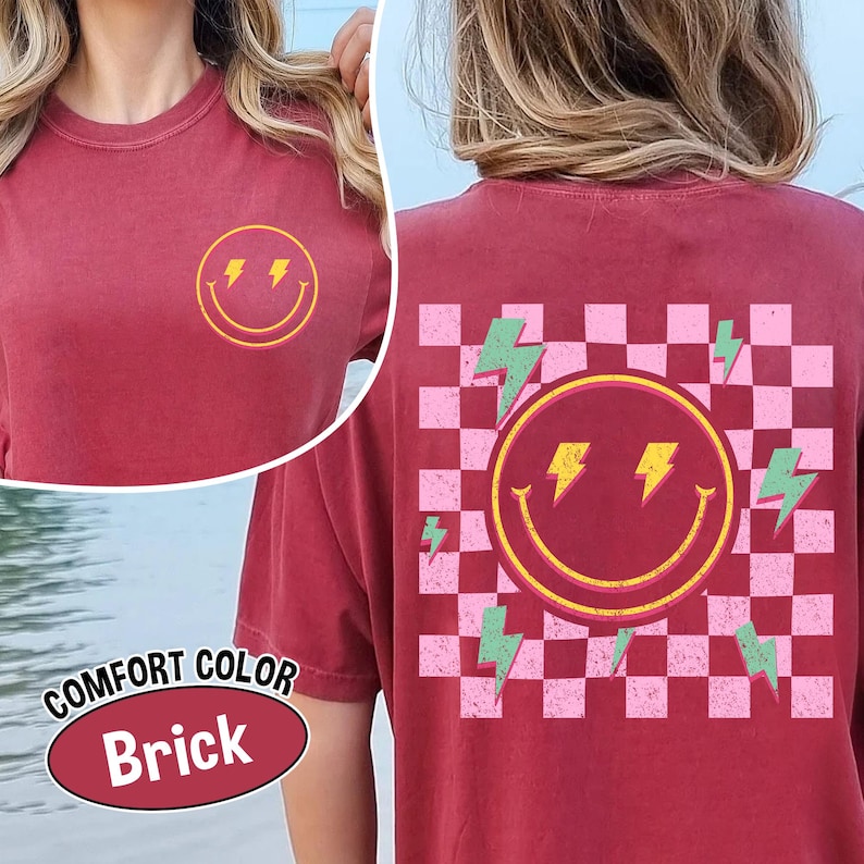 May include: Brick-colored t-shirt with a smiley face and lightning bolt design. The front features a yellow smiley face with lightning bolt eyes. The back has a larger smiley face with a checkered pink and white background and green lightning bolts.