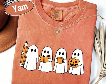 May include: A yam-colored Comfort Colors t-shirt featuring a graphic of four white ghosts, each holding a different item: a pencil, a book, a mug, and a jack-o'-lantern. A gold necklace and a brown hat are also visible.