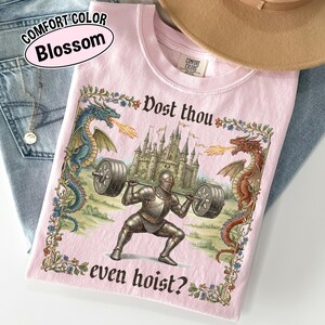 May include: Pink Comfort Colors t-shirt with a medieval knight lifting weights, flanked by dragons. The shirt features the text "Dost thou even hoist?" and "Blossom" in a pink oval.