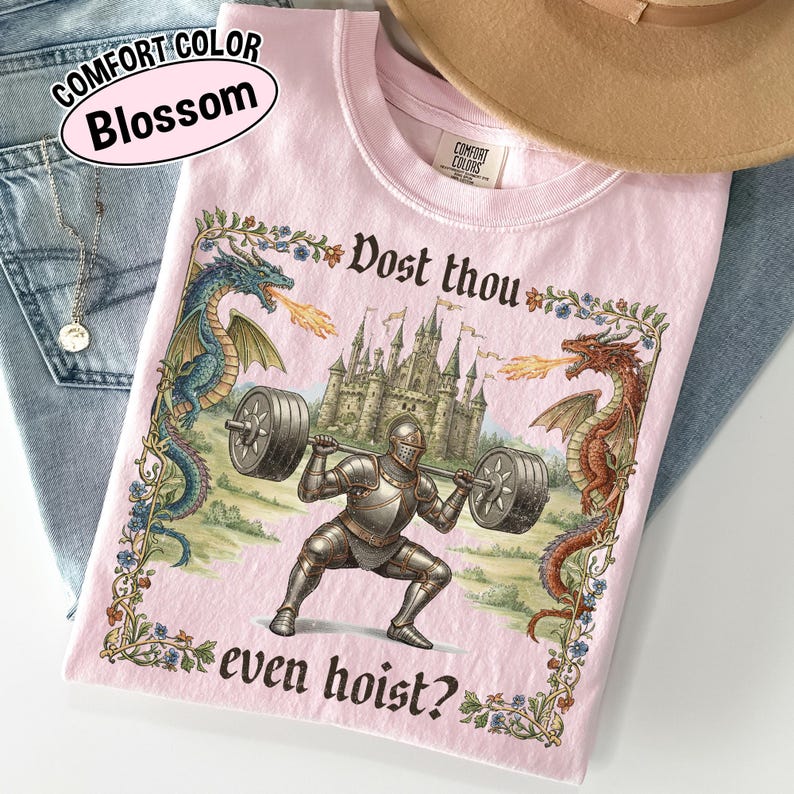 May include: Pink Comfort Colors t-shirt with a medieval knight lifting weights, flanked by dragons. The shirt features the text "Dost thou even hoist?" and "Blossom" in a pink oval.