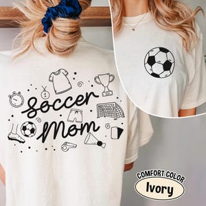 K&ouml;nnte beinhalten: Elfenbeinfarbenes T-Shirt mit einem schwarzen Fu&szlig;ball-Grafik auf der Vorderseite und einem "Soccer Mom"-Design auf der R&uuml;ckseite. Das R&uuml;ckendesign enth&auml;lt fu&szlig;ballbezogene Illustrationen wie eine Troph&auml;e, Shorts und ein Tor.