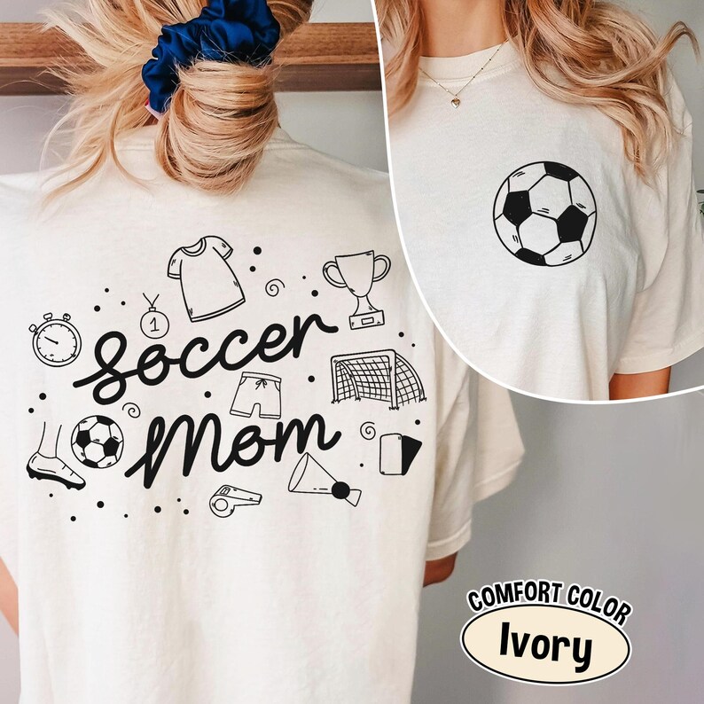 K&ouml;nnte beinhalten: Elfenbeinfarbenes T-Shirt mit einem schwarzen Fu&szlig;ball-Grafik auf der Vorderseite und einem "Soccer Mom"-Design auf der R&uuml;ckseite. Das R&uuml;ckendesign enth&auml;lt fu&szlig;ballbezogene Illustrationen wie eine Troph&auml;e, Shorts und ein Tor.