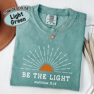 May include: Light green t-shirt with the text "BE THE LIGHT" and "matthew 5:14" in white. The shirt features a sunburst design with an orange sun. A silver necklace with a pendant is on the shirt.