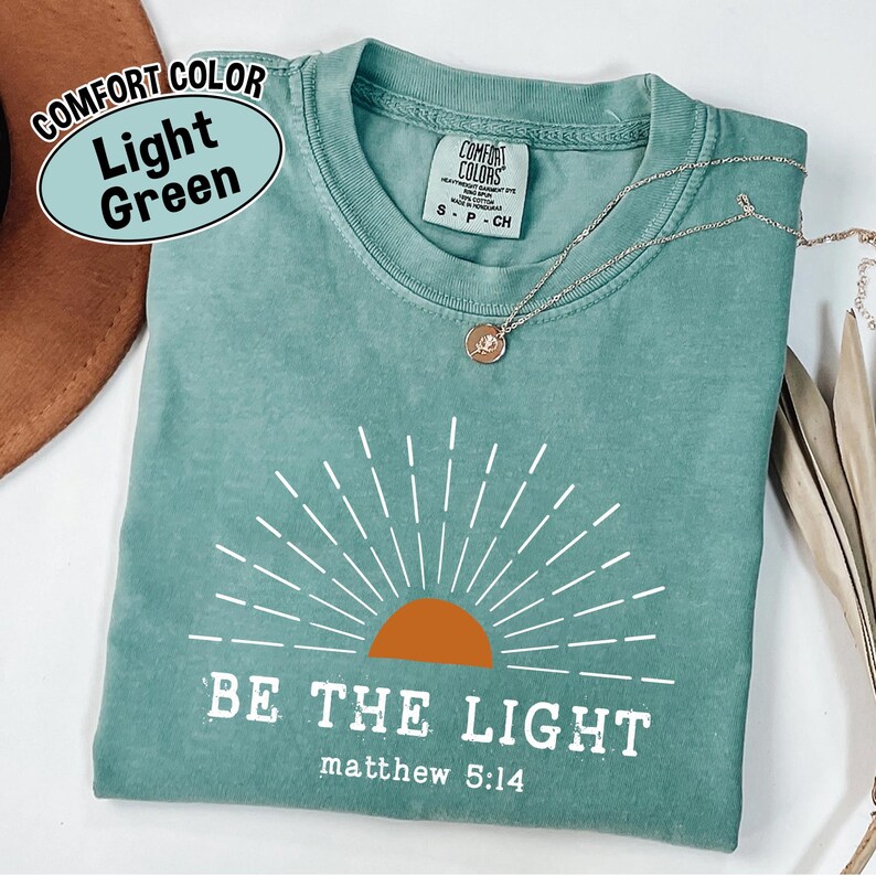 May include: Light green t-shirt with the text "BE THE LIGHT" and "matthew 5:14" in white. The shirt features a sunburst design with an orange sun. A silver necklace with a pendant is on the shirt.