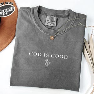 Peut inclure: Un t-shirt gris avec le texte "God is Good" en lettres blanches. Le t-shirt a un petit motif blanc d'une plante avec deux mains qui s'élèvent vers elle.