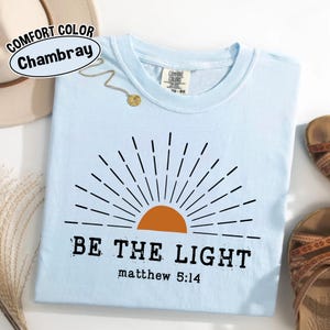 May include: Light blue t-shirt with a graphic of a rising sun in orange and black, and the text "BE THE LIGHT" with "matthew 5:14" below. The shirt is made of a soft material and is a Comfort Color Chambray.