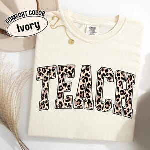 May include: Ivory-colored Comfort Colors t-shirt with the word "TEACH" in a leopard print design. A gold necklace with a pendant is draped on the shirt. The shirt has a crew neck and short sleeves.