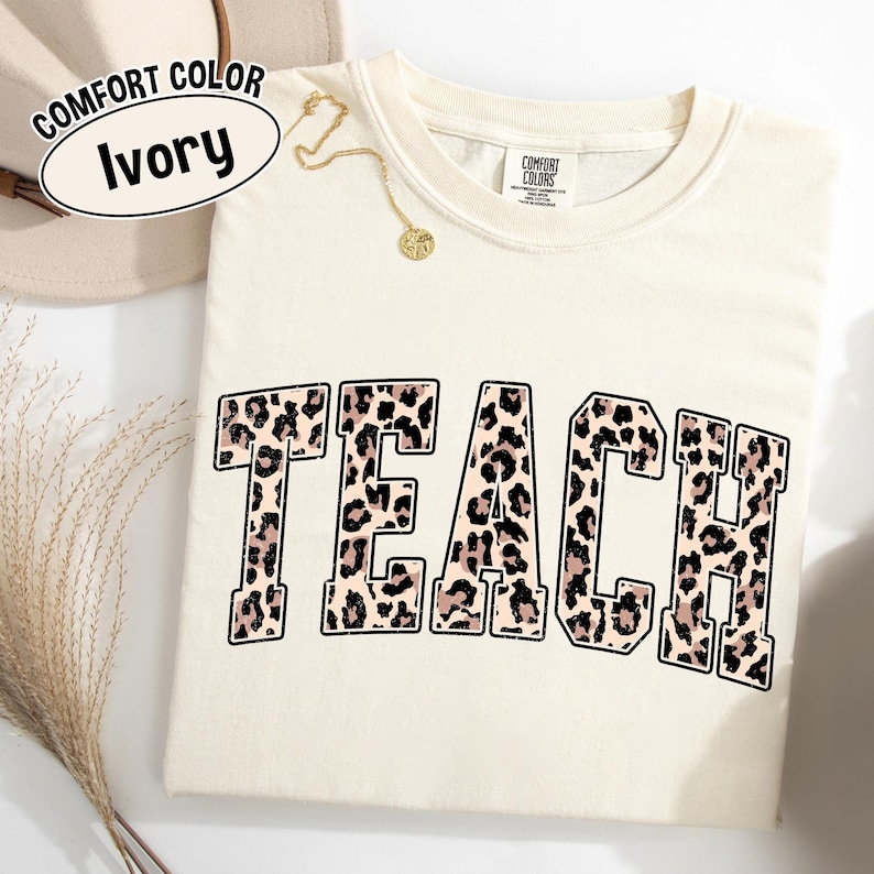 May include: Ivory-colored Comfort Colors t-shirt with the word "TEACH" in a leopard print design. A gold necklace with a pendant is draped on the shirt. The shirt has a crew neck and short sleeves.