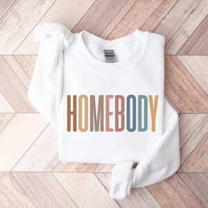 May include: A white sweatshirt with the word "HOMEBODY" printed across the front in a colorful, retro font. The sweatshirt is folded neatly on a wooden surface.