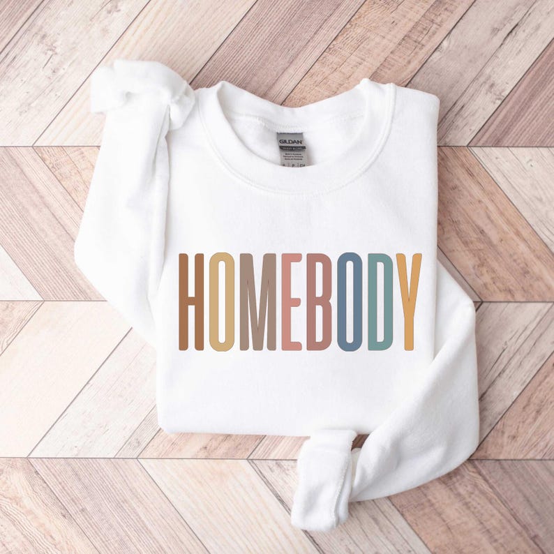 May include: A white sweatshirt with the word "HOMEBODY" printed across the front in a colorful, retro font. The sweatshirt is folded neatly on a wooden surface.