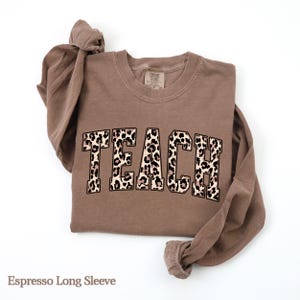 May include: Espresso-colored long-sleeve shirt with the word "TEACH" in a leopard print design across the front. The shirt is folded, with the sleeves knotted at the top. The shirt is made of a soft material.
