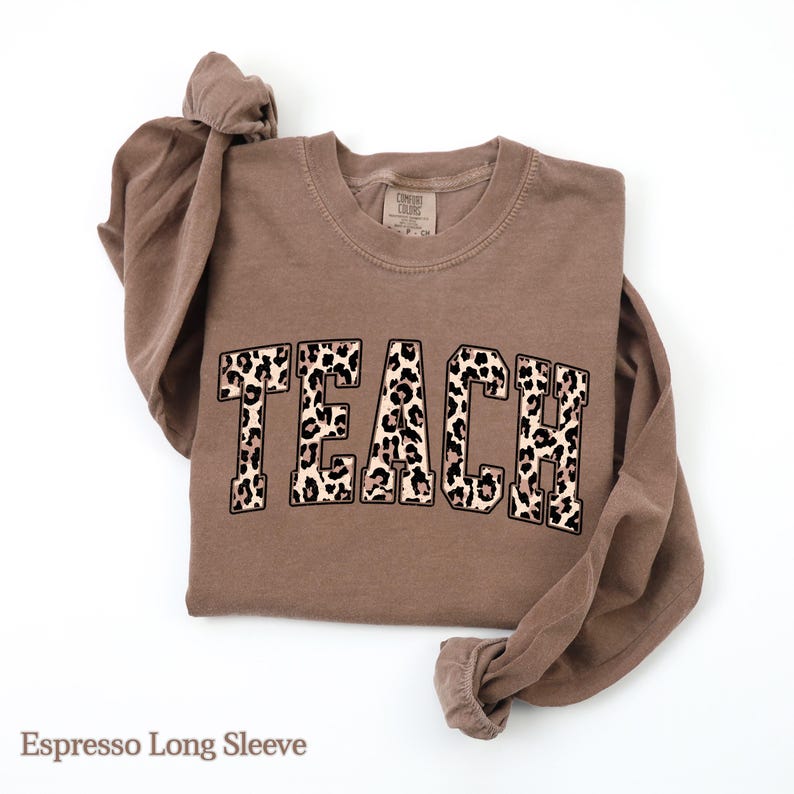 May include: Espresso-colored long-sleeve shirt with the word "TEACH" in a leopard print design across the front. The shirt is folded, with the sleeves knotted at the top. The shirt is made of a soft material.