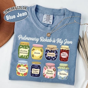 May include: Blue jean Comfort Colors t-shirt with the phrase "Pulmonary Rehab is My Jam." The shirt features illustrations of jam jars with labels like "Breathing Retraining" and "Nutrition." A gold necklace is draped across the shirt.