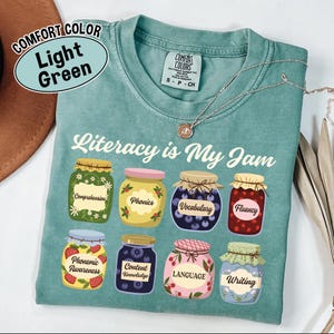 May include: Light green t-shirt with the phrase "Literacy is My Jam" and illustrations of jam jars labeled with literacy terms like "Phonics" and "Vocabulary". A silver necklace is draped across the shirt.