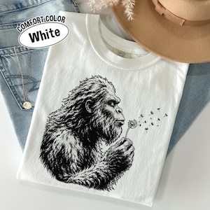 Puede incluir: Una camiseta blanca de cuello redondo con una ilustraci&oacute;n gr&aacute;fica en negro de una criatura Bigfoot soplando un diente de le&oacute;n. La prenda incluye una etiqueta Comfort Colors en el cuello y un texto en la esquina superior izquierda que dice COMFORT COLOR White.
