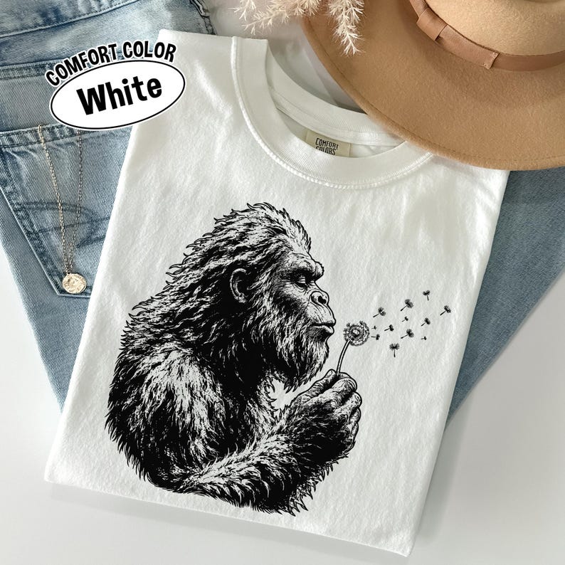 Puede incluir: Una camiseta blanca de cuello redondo con una ilustraci&oacute;n gr&aacute;fica en negro de una criatura Bigfoot soplando un diente de le&oacute;n. La prenda incluye una etiqueta Comfort Colors en el cuello y un texto en la esquina superior izquierda que dice COMFORT COLOR White.