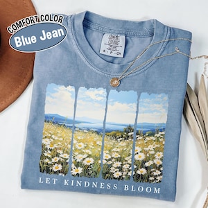 May include: A blue Comfort Colors t-shirt with a landscape graphic of a field of daisies and the text "LET KINDNESS BLOOM." The shirt has a crew neck and short sleeves. A gold necklace with a pendant is draped across the shirt.