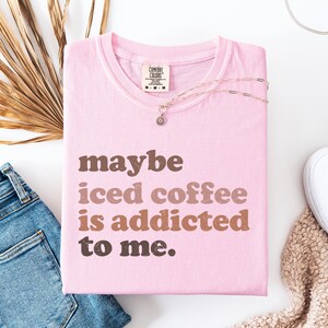 Peut inclure: T-shirt rose clair Comfort Colors avec l'inscription "maybe iced coffee is addicted to me" en lettres marron et beige. Un collier argent&eacute; avec un petit pendentif est pos&eacute; sur le t-shirt. Un jean bleu et une chaussure blanche sont visibles.
