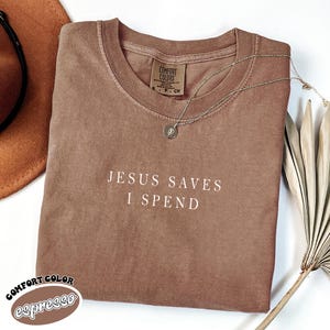 May include: A brown t-shirt with the text "Jesus Saves I Spend" in white lettering. The shirt is made of a soft, comfortable material and has a distressed look. The shirt is folded and laid flat on a white surface.