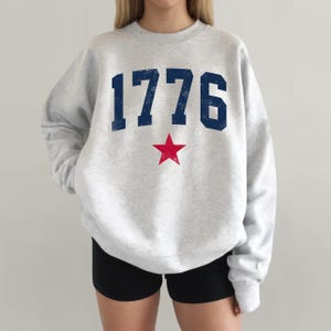 May include: A light gray sweatshirt with the number "1776" in navy blue and a red star. The sweatshirt is a crew neck style.
