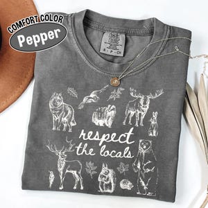 May include: A grey Comfort Colors t-shirt with a white graphic of various animals and the text "respect the locals." The shirt has a crew neck and short sleeves. A silver necklace with a pendant is draped across the shirt.