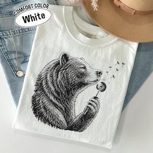 May include: A white t-shirt featuring a black ink-style illustration of a bear holding a dandelion. The bear is shown in profile, blowing on the dandelion seeds. The shirt has a crew neckline and a Comfort Colors label on the inner collar.