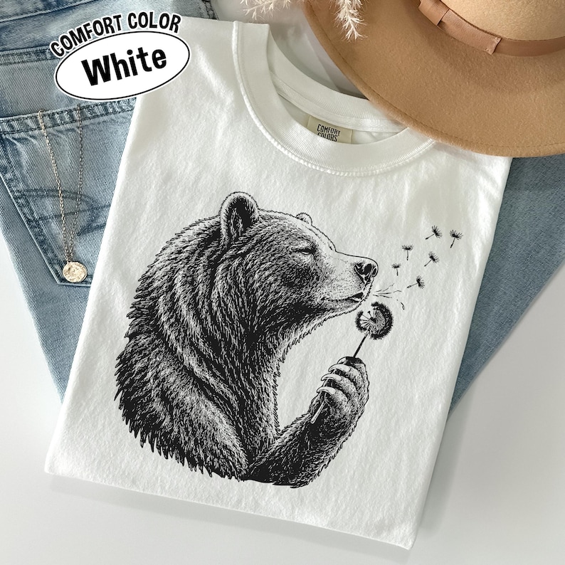 May include: A white t-shirt featuring a black ink-style illustration of a bear holding a dandelion. The bear is shown in profile, blowing on the dandelion seeds. The shirt has a crew neckline and a Comfort Colors label on the inner collar.