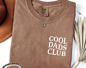 May include: A brown t-shirt with the text "COOL DADS CLUB" printed in white. The shirt is made of a soft, comfortable material and has a relaxed fit.