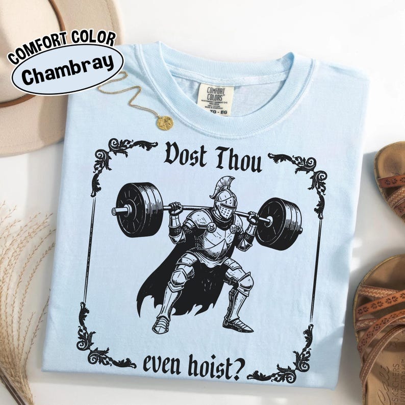 Może przedstawiać: Jasnoniebieska koszulka z czarną grafiką rycerza podnoszącego sztangę z napisem "Dost Thou even hoist?". Koszulka jest oznaczona jako "Comfort Color Chambray". Widoczny jest złoty naszyjnik i sandały.