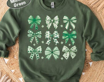 May include: Military green sweatshirt featuring nine green bows with white accents and shamrock details. The bows vary in design, with some having plaid patterns. The sweatshirt is a crew neck style.