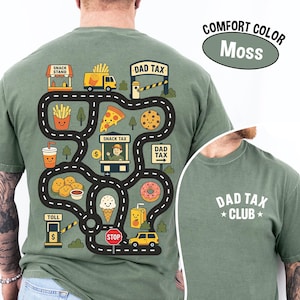 May include: Moss green t-shirt with a playful road map design featuring snack stands, food trucks, and toll booths. The graphic includes the text "DAD TAX" and "SNACK TAX". The back of the shirt has the text "DAD TAX CLUB".