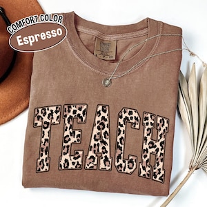 May include: A brown Comfort Colors Espresso t-shirt with the word "TEACH" in large leopard print letters. A silver necklace with a pendant is draped across the shirt. A brown hat is visible in the upper left corner.