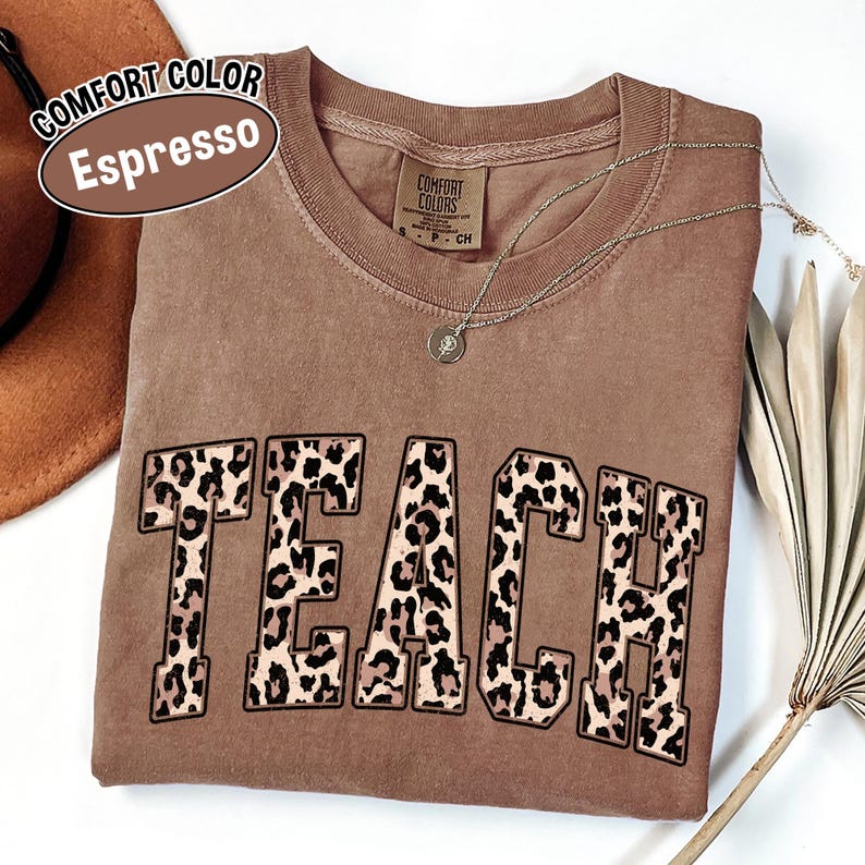 May include: A brown Comfort Colors Espresso t-shirt with the word "TEACH" in large leopard print letters. A silver necklace with a pendant is draped across the shirt. A brown hat is visible in the upper left corner.