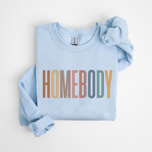 May include: A light blue sweatshirt with the word "HOMEBODY" printed across the front in a colorful, retro font. The long sleeves are folded, and the sweatshirt is laid flat on a white surface. The Gildan brand tag is visible.