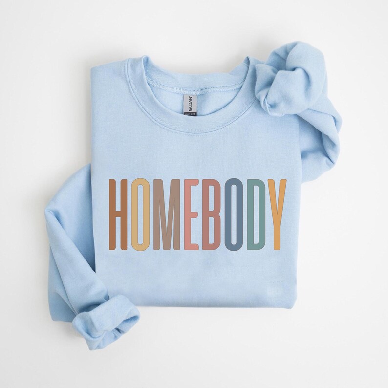 May include: A light blue sweatshirt with the word "HOMEBODY" printed across the front in a colorful, retro font. The long sleeves are folded, and the sweatshirt is laid flat on a white surface. The Gildan brand tag is visible.