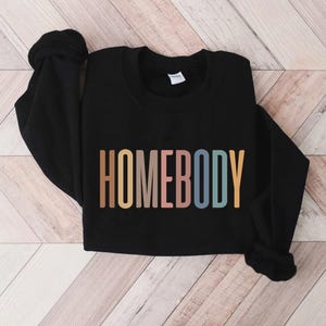May include: Black sweatshirt with the word "HOMEBODY" in a colorful, retro font across the front. The text is in shades of brown, pink, blue, and yellow. The sweatshirt is lying on a light wood-grain surface.
