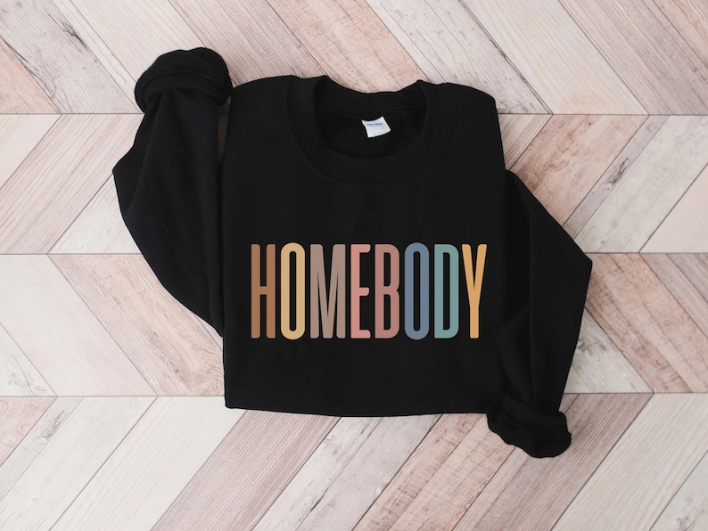 May include: Black sweatshirt with the word "HOMEBODY" in a colorful, retro font across the front. The text is in shades of brown, pink, blue, and yellow. The sweatshirt is lying on a light wood-grain surface.