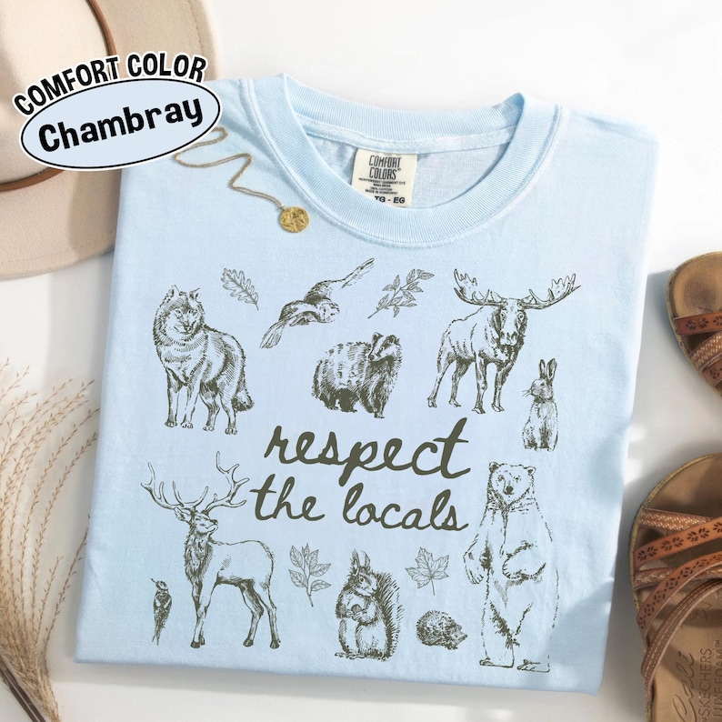 May include: Pale blue Comfort Colors Chambray t-shirt featuring a graphic of woodland creatures and the text "respect the locals". The top has a crew neck and short sleeves. A gold necklace and sandals are visible.