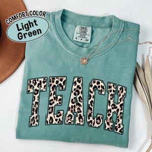 May include: Light green Comfort Colors t-shirt with the word "TEACH" in a leopard print design. A gold necklace with a pendant is on the shirt. The shirt is folded and placed on a light-colored surface.