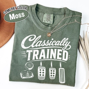 May include: A moss green short-sleeve t-shirt with a crew neckline. The front features white screen-printed text that reads Classically TRAINED above a graphic of a manual gear shifter and three car pedals. The shirt has a Comfort Colors label.