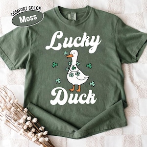 Puede incluir: Una camiseta verde musgo con las palabras "Lucky Duck" en escritura blanca. En el centro hay un pato de dibujos animados con tr&eacute;boles. La camiseta tambi&eacute;n tiene las palabras "Comfort Color" y "Moss" en un &oacute;valo blanco.