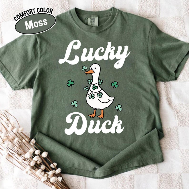 Puede incluir: Una camiseta verde musgo con las palabras "Lucky Duck" en escritura blanca. En el centro hay un pato de dibujos animados con tr&eacute;boles. La camiseta tambi&eacute;n tiene las palabras "Comfort Color" y "Moss" en un &oacute;valo blanco.