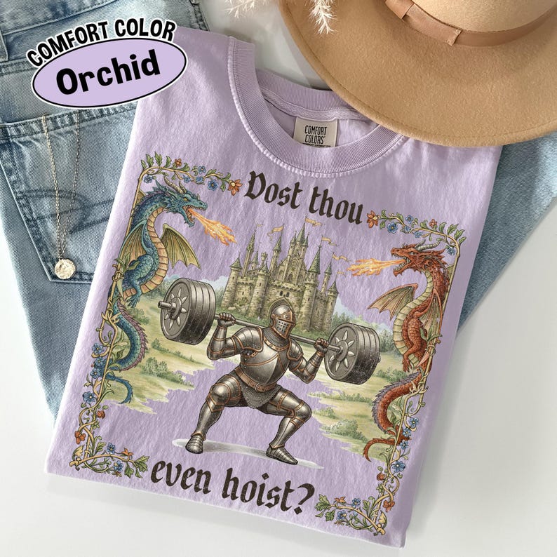 May include: Orchid-colored t-shirt with a medieval knight lifting weights, flanked by dragons. The shirt features the text "Dost thou even hoist?" and "Comfort Colors" branding. A tan hat and blue jeans are in the background.