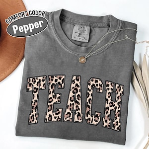 May include: A gray Comfort Colors t-shirt with the word "TEACH" in large, leopard print letters. The shirt has a crew neck and short sleeves. A silver necklace with a small pendant is draped on the shirt.
