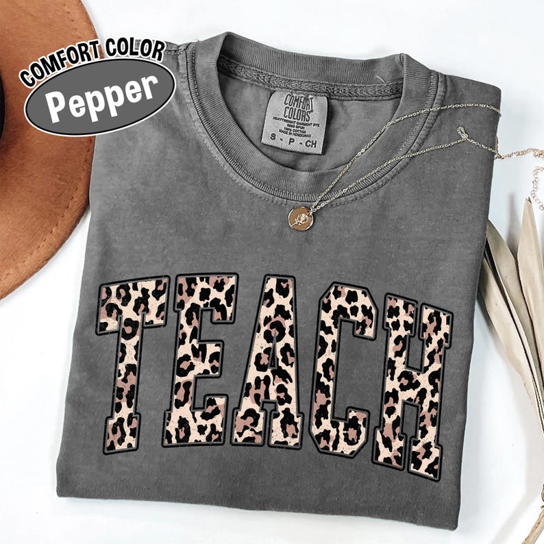 May include: A gray Comfort Colors t-shirt with the word "TEACH" in large, leopard print letters. The shirt has a crew neck and short sleeves. A silver necklace with a small pendant is draped on the shirt.